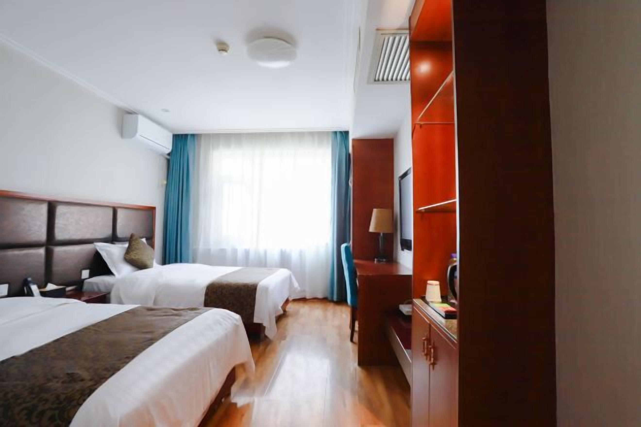 A hotel room with two beds, a wooden floor, a desk with a chair, and a window with curtains.