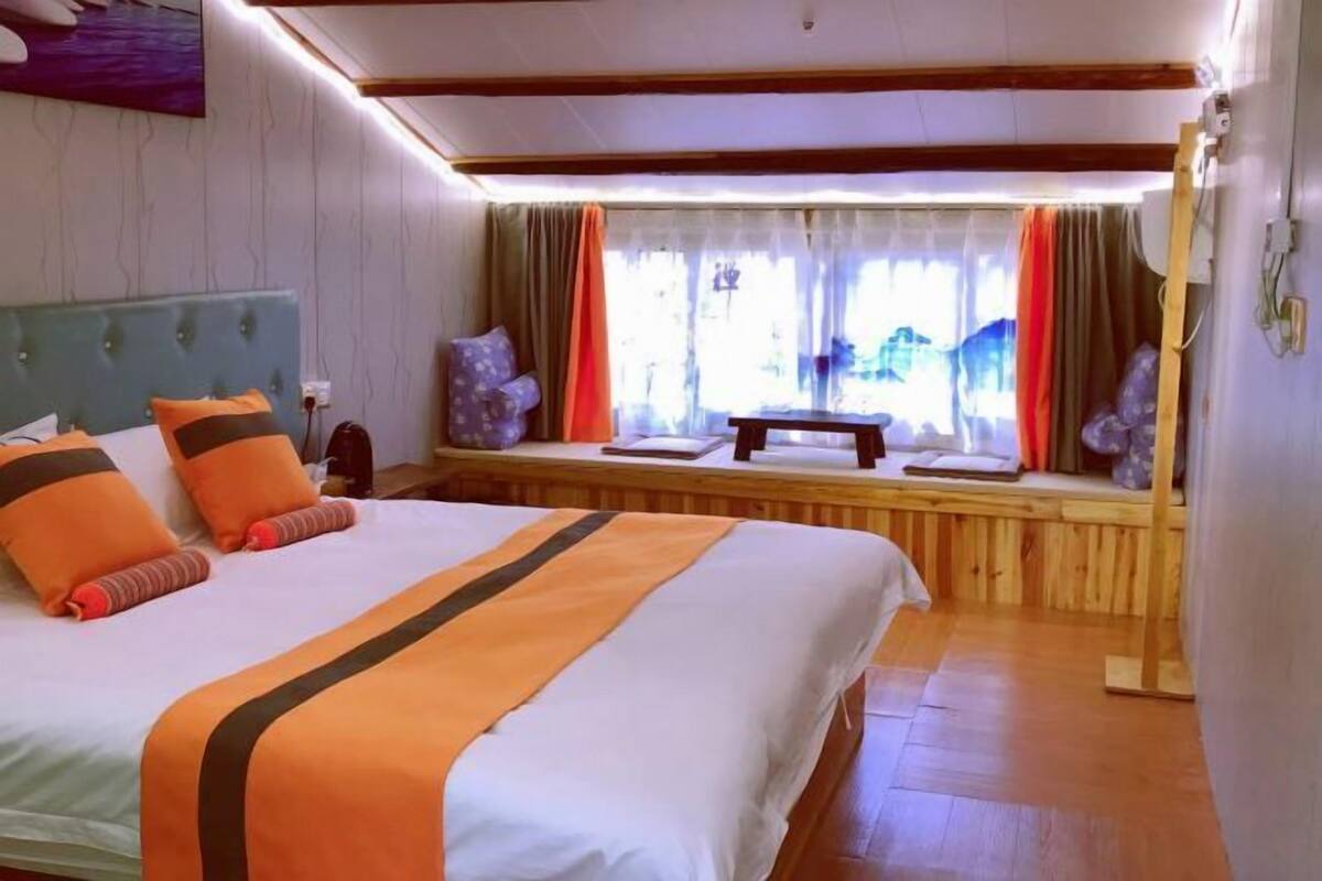 Ein Hotelzimmer mit einem großen Bett, einer hölzernen Bank, einem fenster mit Vorhängen und Blick ins Freie.