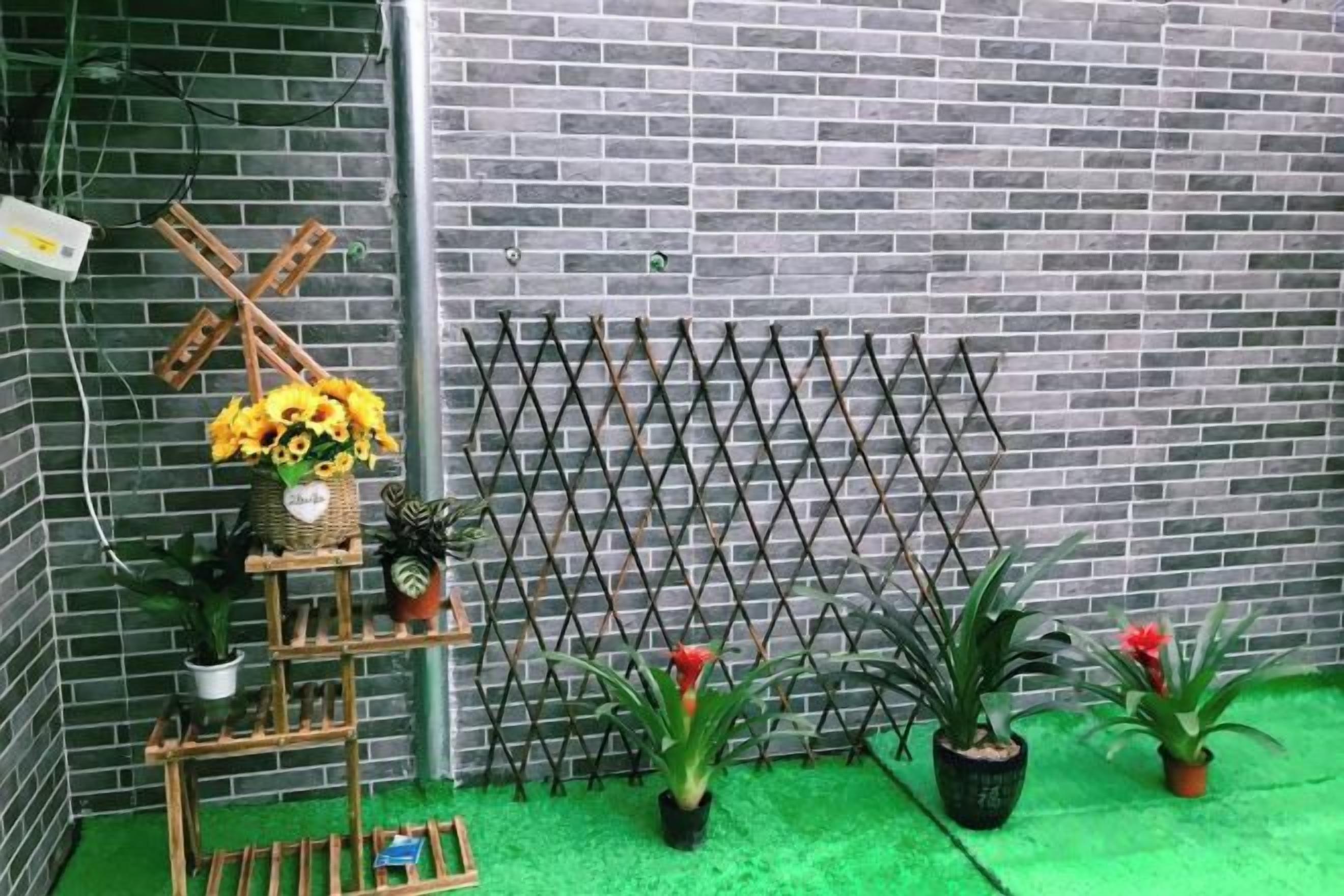 Un muro de ladrillo con una cerca metálica, un soporte de madera con flores y plantas en macetas sobre una superficie verde.