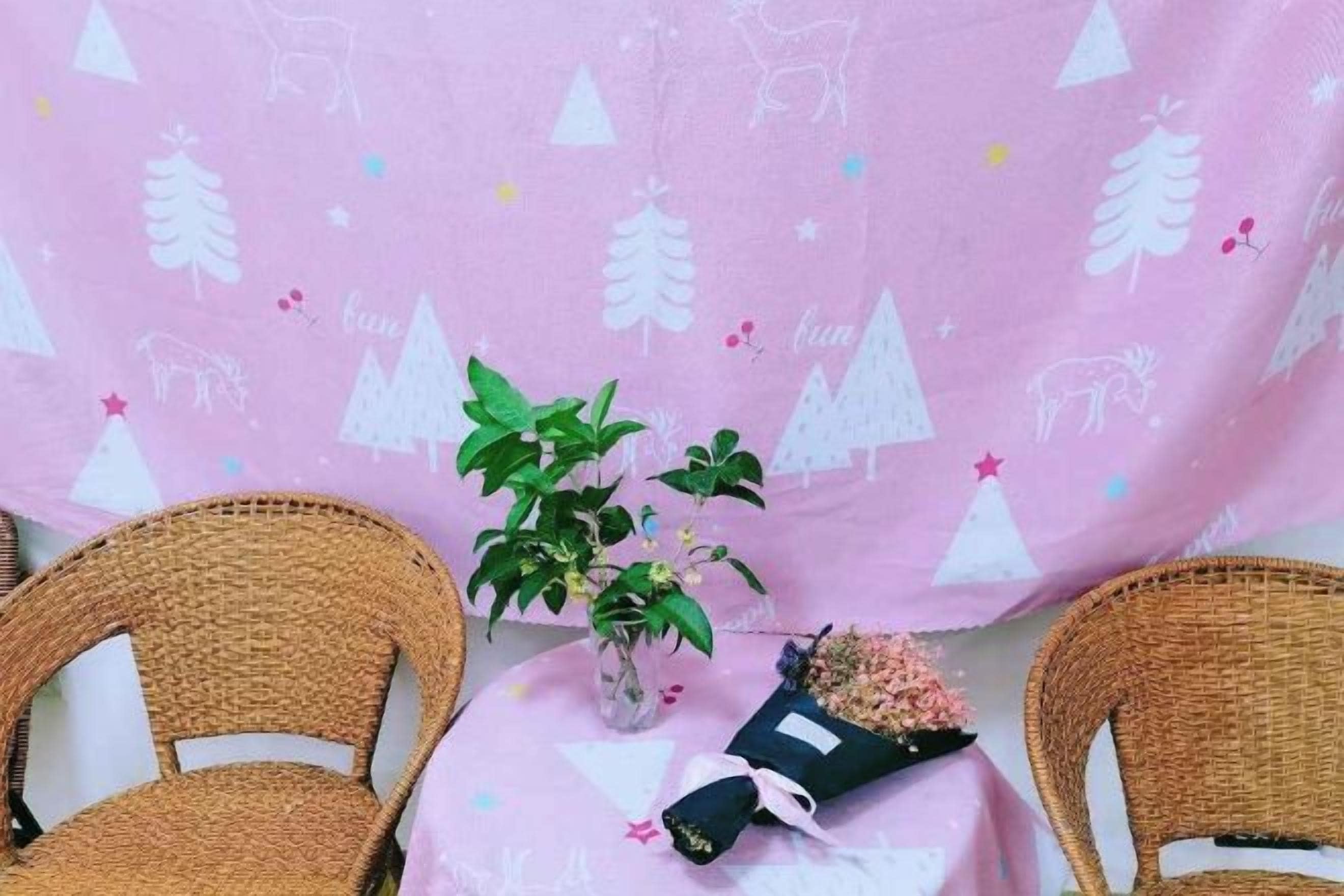Una habitación con mantel rosa, dos sillas de mimbre, una planta en maceta y un ramo de flores.