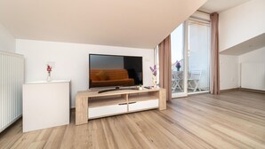 Una sala de estar moderna con una televisión de pantalla plana, piso de madera, un sofá, una mesita auxiliar blanca pequeña y un cerramiento de vidrio corredizo que da a un espacio exterior.