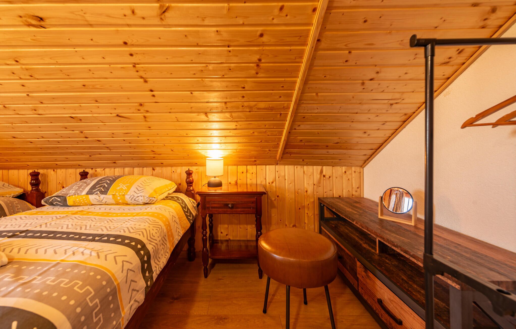 Habitació acollidora amb parets i sostre de fusta, un llit amb roba de llit estampada, una taula petita amb una làmpada, un tamboret i un mirall a la paret.