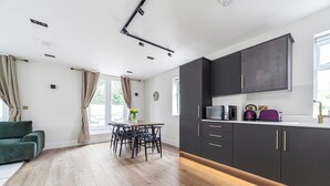 A modern kitchen with dark cabinets, a dining area with a wooden table and chairs, and a green sofa in a bright living space.
