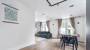 A spacious living area with a wooden floor, a dining table with chairs, a sofa, and a large window with curtains.