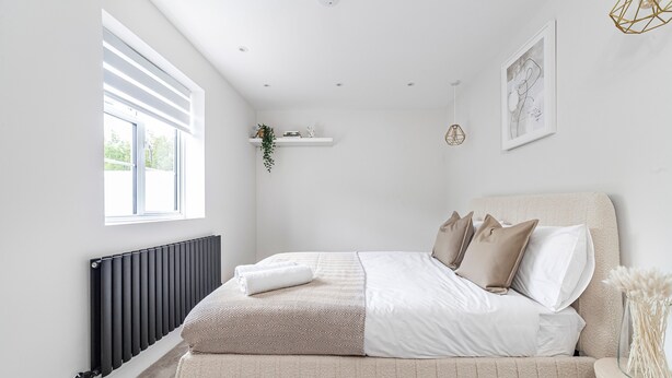 A bedroom with a bed, a radiator, a window, a shelf with a plant, and a framed picture on the wall.