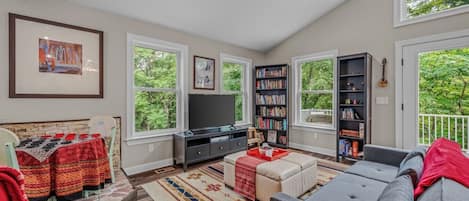 Uma sala de estar ampla, com uma janela grande, televisão, estante de livros e um sofá.