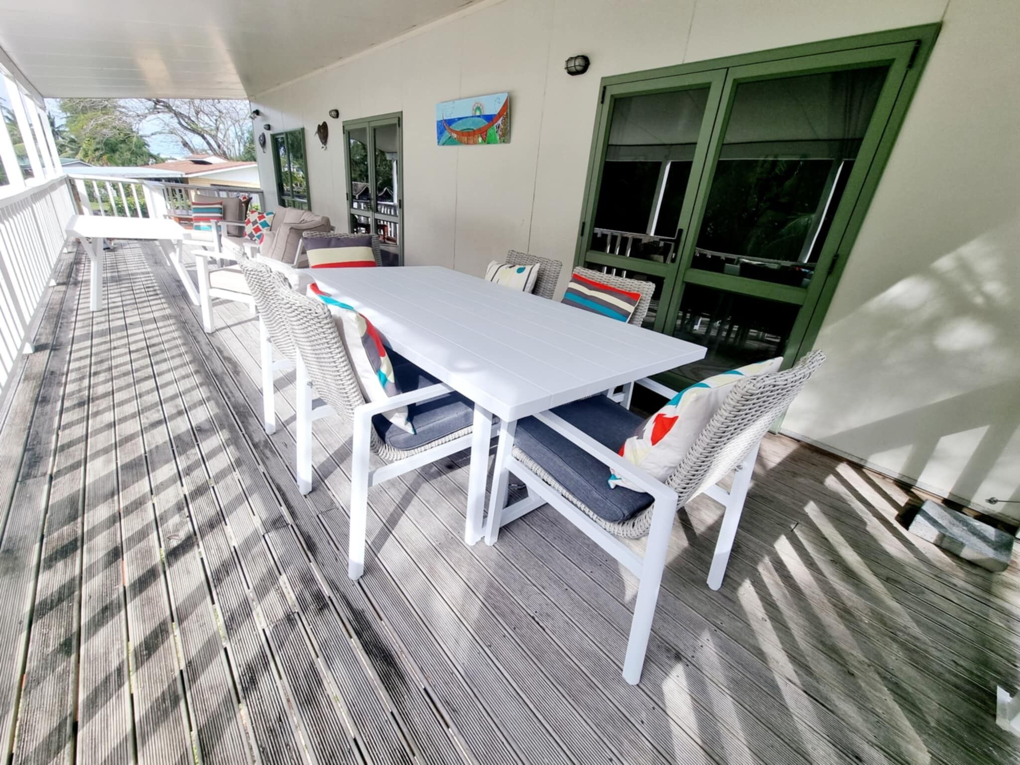 A covered outdoor dining area with a white table and wicker chairs, a wooden deck, and a green-framed window.