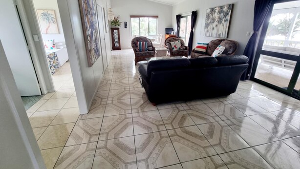 A spacious living room with a large black sofa, a dining area with a table and chairs, and a kitchen in the background. - Natura Nikao Beach Villa (Rarotonga)