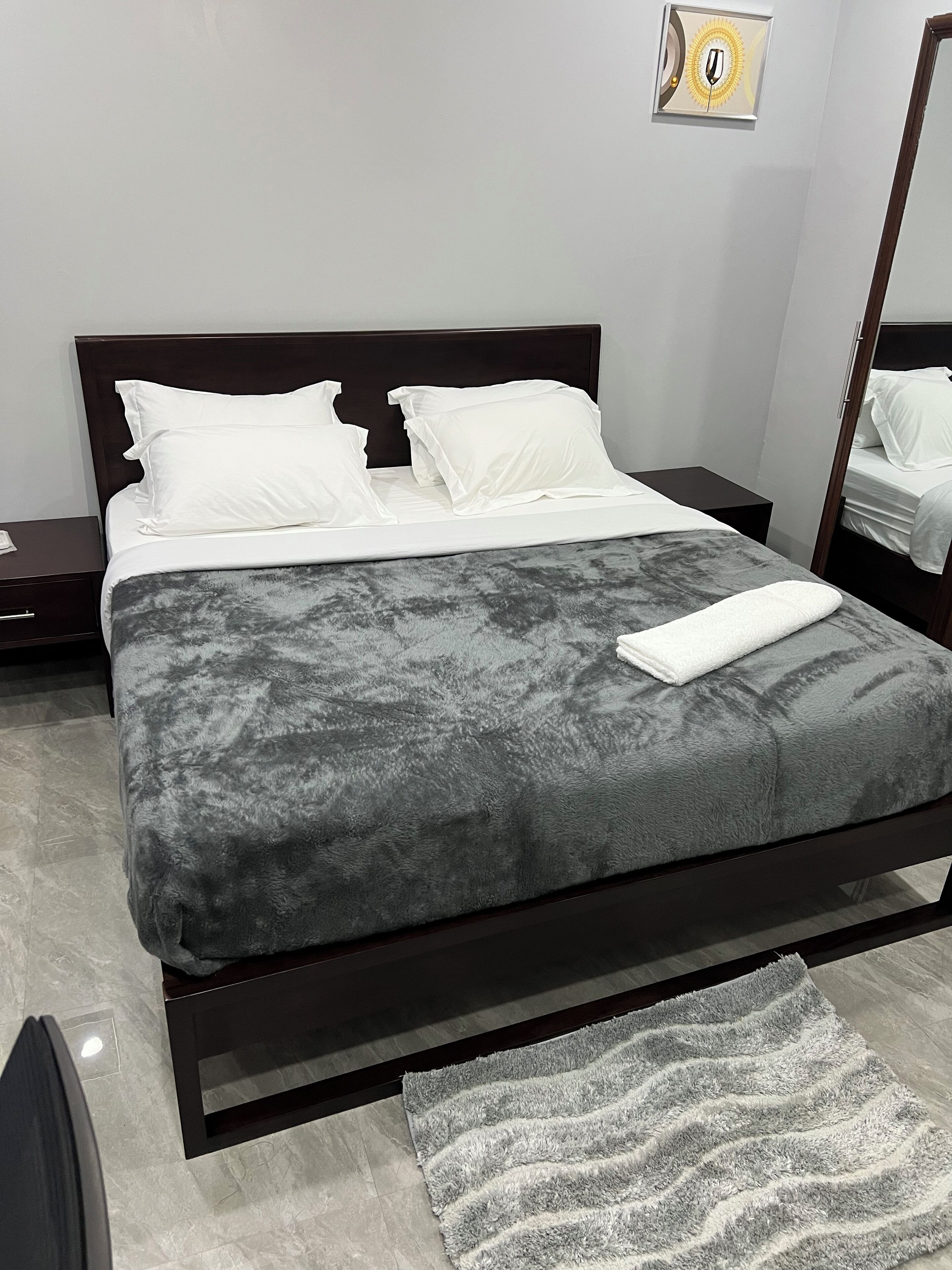 Una cama bien hecha con un edredón gris, almohadas blancas y una toalla blanca doblada. Hay una alfombra pequeña en el suelo y una mesita de noche con una lámpara junto a la cama.