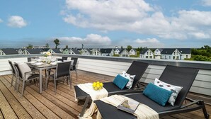 A rooftop terrace with a dining table set, lounge chairs, and a view of white houses in the background.