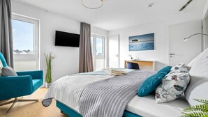 A modern hotel room with a large bed, a desk, a TV, and a view of the sea.