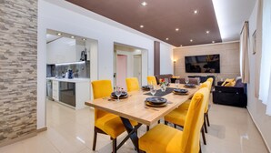 Un comedor moderno con una mesa de madera y sillas amarillas, una cocina al fondo y un televisor montado en la pared.