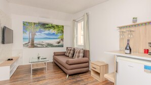 Ein gemütliches Wohnzimmer mit einer braunen Couch, einem Glastisch, einem Gemälde mit Strandmotiv, einem kleinen Holznachttisch und einer weißen Kommode mit einer Weinflasche und Gläsern.