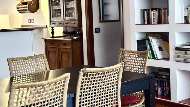 Zona pranzo con sedie in vimini, tavolo in legno, un mobiletto con ante in vetro e una libreria piena di libri.
