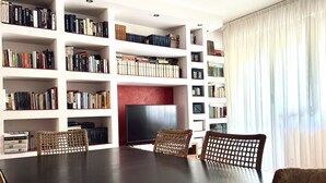 Una stanza con un grande tavolo di legno scuro, sedie in vimini, un televisore a schermo piatto e scaffali pieni di libri.