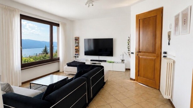 A modern living room with a large window, a flat-screen TV, a sofa, and a door leading to another room. - Cà Sulà, large detached house with garden and beautiful lake view (Maccagno con Pino e Veddasca)