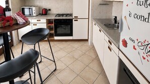 A modern kitchen with white cabinets, a gas stove, a microwave, and a refrigerator. There are two black bar stools and a small dining table. - Cà Sulà, large detached house with garden and beautiful lake view (Maccagno con Pino e Veddasca)
