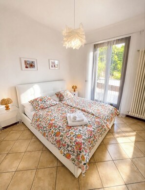 A bedroom with a bed, two bedside tables, a lamp, a window with curtains, and a radiator.