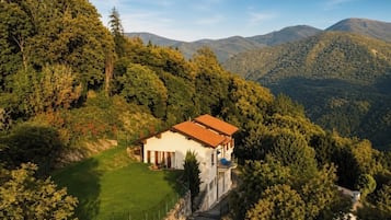 Une maison de plain-pied avec un toit recouvert de tuiles rouges, entourée d’une végétation luxuriante et avec des collines en arrière-plan.