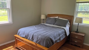 A bedroom with a wooden bed, two bedside tables, and a view of greenery through the windows.