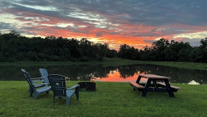 A picnic setup with chairs and a table by a calm lake, surrounded by trees under a sunset sky.