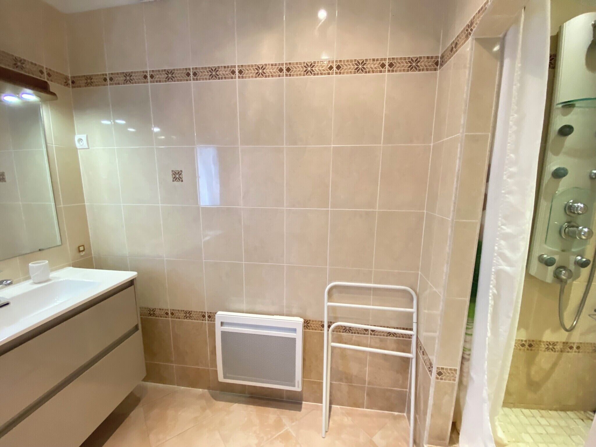 Une salle de bain avec un lavabo blanc, un porte-serviettes, une douche dotée d’une porte en verre et une grille d’aération sur le mur.