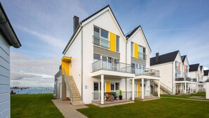 Un immeuble résidentiel moderne à plusieurs étages, doté de balcons, d’un porche couvert et offrant une vue sur l’eau.