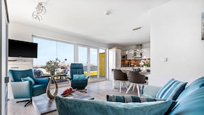 Un salon moderne comprenant un canapé, des fauteuils, une table basse et un téléviseur. De grandes fenêtres offrent une vue sur la mer.