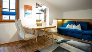 A modern living room with a wooden dining table, white chairs, a blue sofa, and a large window providing natural light.