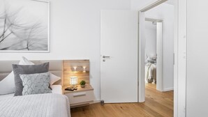 Quarto com cama, mesa de cabeceira, abajur e porta que dá para outro cômodo.