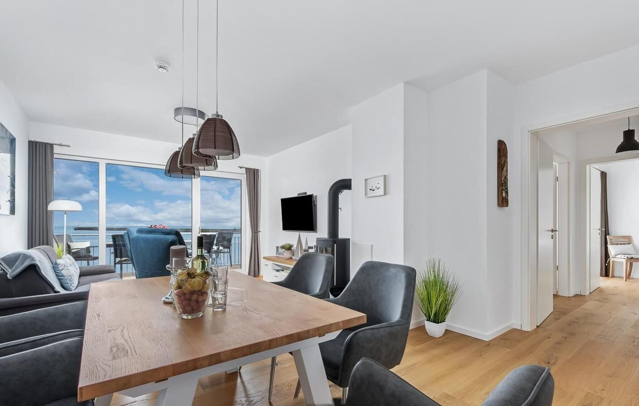 A modern living room with a wooden dining table, grey sofas, and a large window offering a view of the sea.