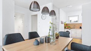 Uma sala de jantar moderna com uma mesa de madeira, cadeiras pretas e três luminárias pendentes. Uma cozinha com armários brancos e uma pia é visível ao fundo.