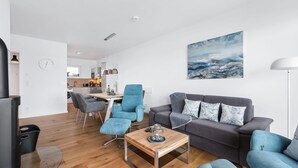 Uma sala de estar moderna com um sofá cinzento, poltronas azuis, uma mesa de centro de madeira e uma cozinha ao fundo.