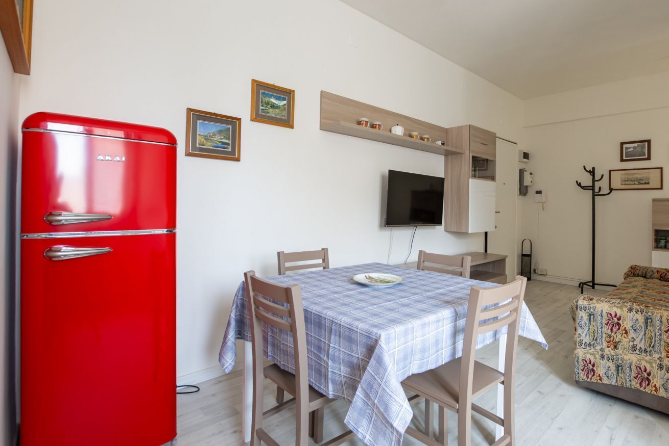 A compact living space with a red refrigerator, a dining table with four chairs, a television mounted on the wall, and a bed with a floral bedspread.