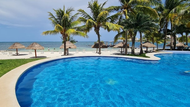 A swimming pool with a curved edge, surrounded by palm trees and beach umbrellas. - Oceanfront Condo at Unique Location (Cancun)