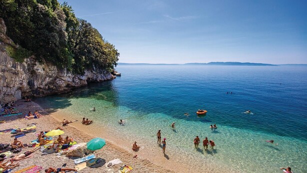 Ein Strand mit klarem Wasser, Menschen, die sich sonnen, und einer felsigen Klippe.