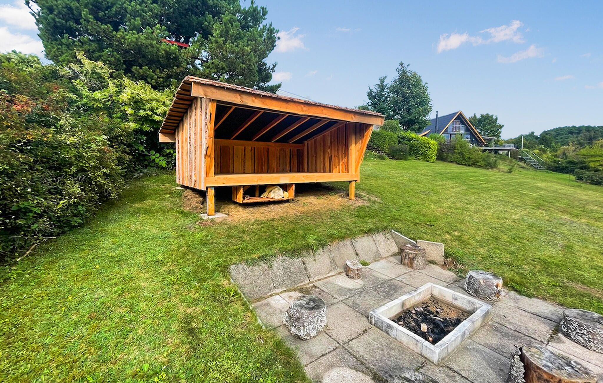 Une structure extérieure en bois, dotée d’un toit incliné, qui pourrait être un abri de jardin ou une petite cabane, située sur une pelouse verdoyante, avec un foyer en pierre à proximité.