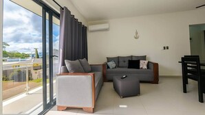 A modern living room with a grey sofa, a small ottoman, and a dining table with chairs. There is a large window with curtains, an air conditioning unit on the wall, and a view of a building outside. - Spacious Apartment w/ Secure Private Parking (Puerto Plata)