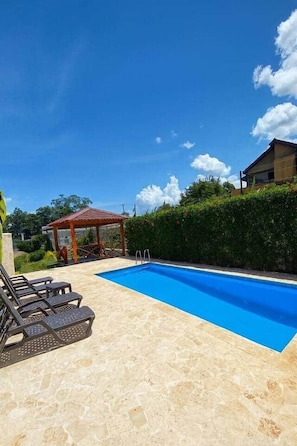 Une piscine avec des chaises longues, un kiosque et une haie.