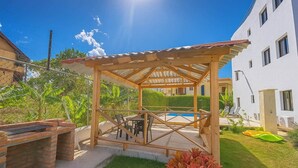 A wooden gazebo structure with a pool in the background, surrounded by greenery and a white building. - Spacious Apartment w/ Secure Private Parking (Puerto Plata)