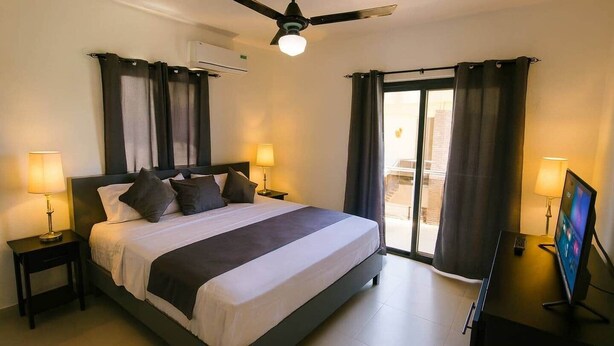 A hotel room with a bed, two bedside tables with lamps, a ceiling fan, a TV, and a sliding glass door to the outside. - Spacious Apartment w/ Secure Private Parking (Puerto Plata)