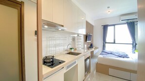 A compact hotel room with a kitchenette, a bed, a window with curtains, and a built-in air conditioning unit. - Warm And Homey Studio At Sky House Alam Sutera Apartment (Tangerang)
