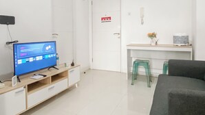 A modern, well-lit room with a flat-screen TV on a wooden TV stand, a desk with a chair, a green stool, and a sofa.