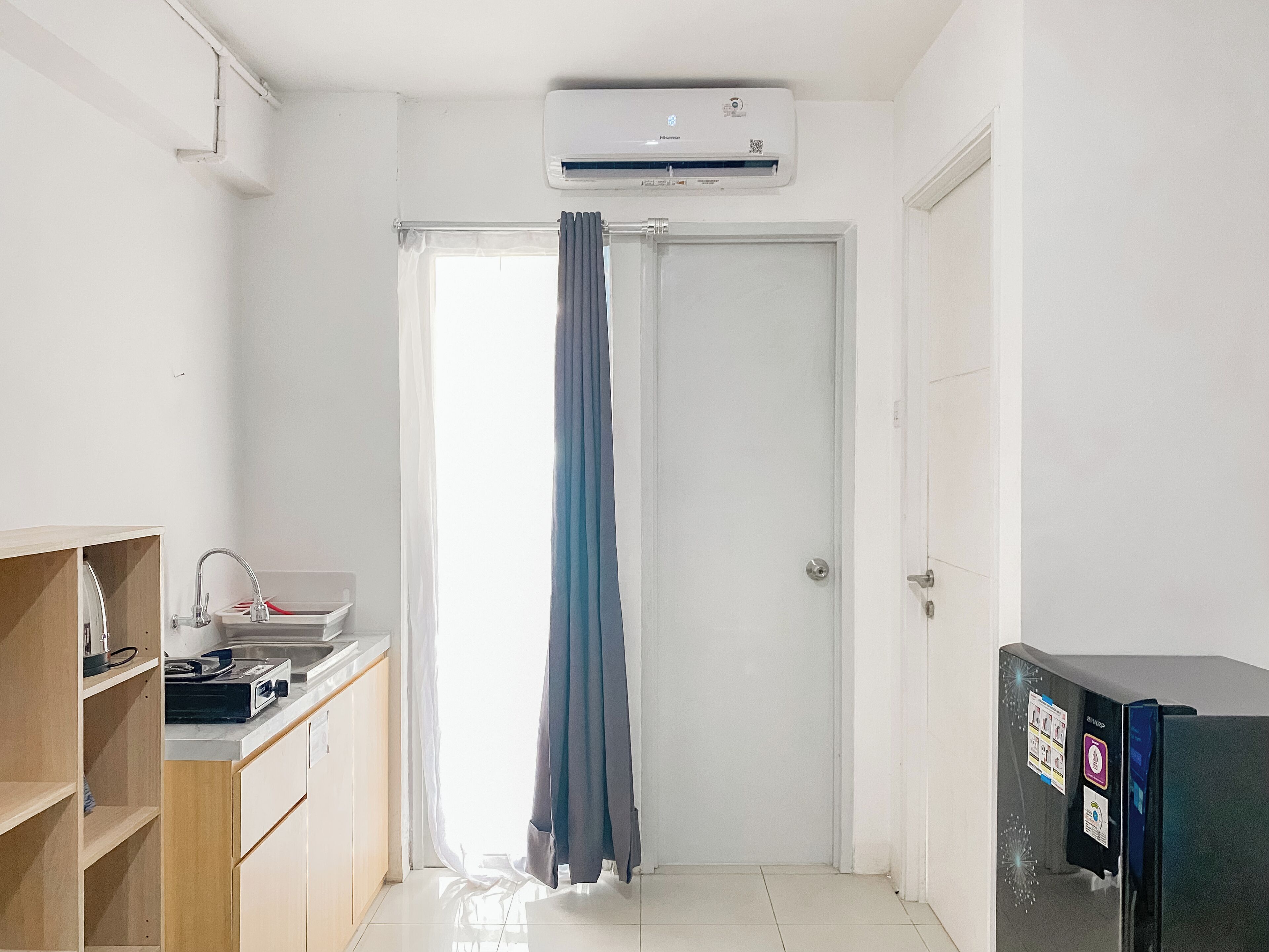 Quarto pequeno e limpo com porta branca, janela com cortina, cozinha compacta com pia e bancada pequena, e unidade de ar condicionado na parede.