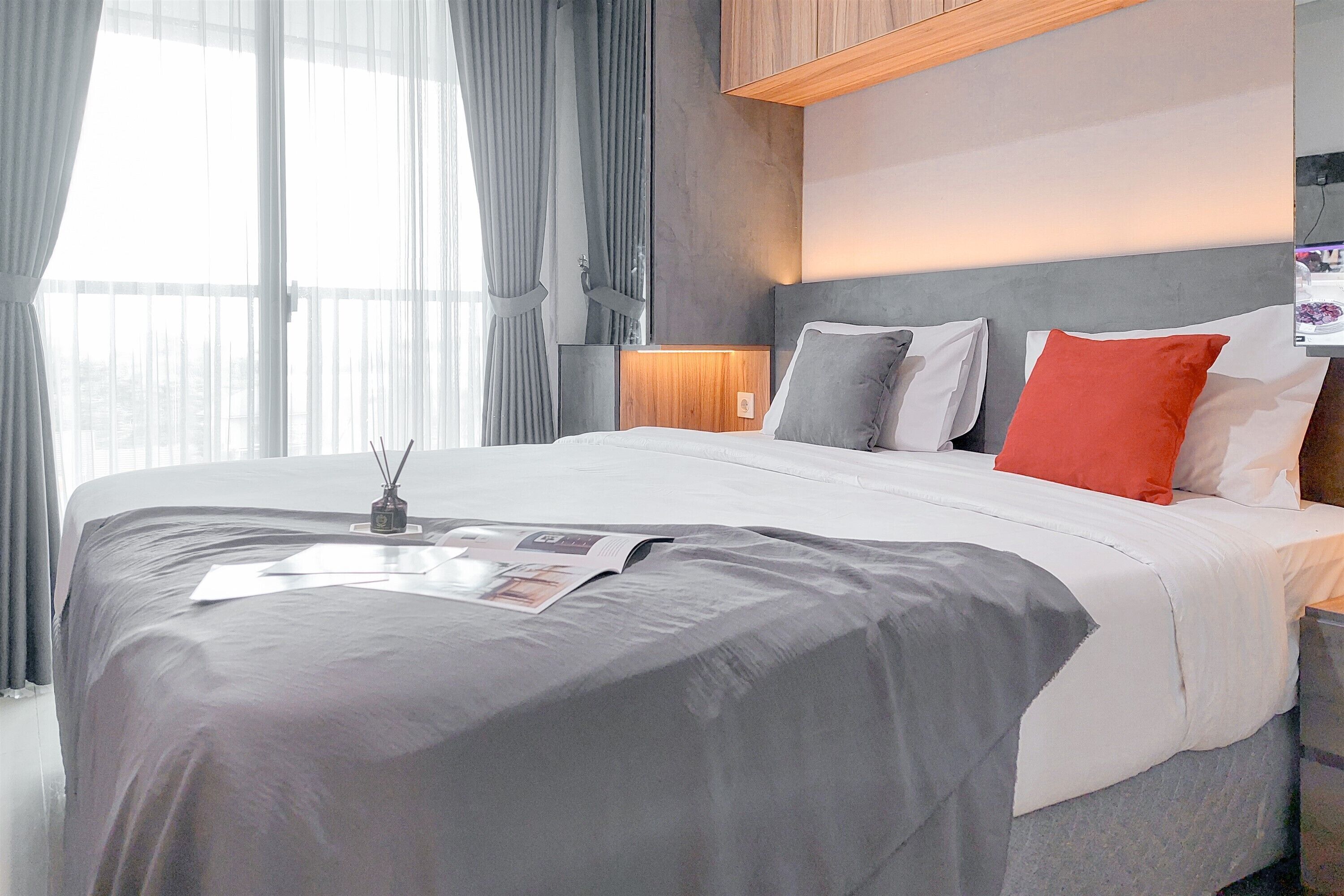 A modern hotel room with a large bed, grey and white bedding, a wooden headboard, and a bedside table with a lamp.