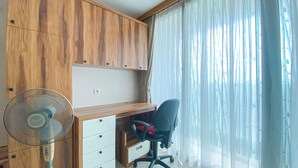 Una stanza con una scrivania in legno, una sedia, un ventilatore e una finestra con tende trasparenti.