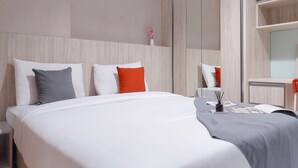 Una cama bien hecha con ropa de cama blanca y gris, un plaid gris y dos almohadas (una gris y una roja). Una mesita de noche con una revista y un objeto negro está al lado de la cama.