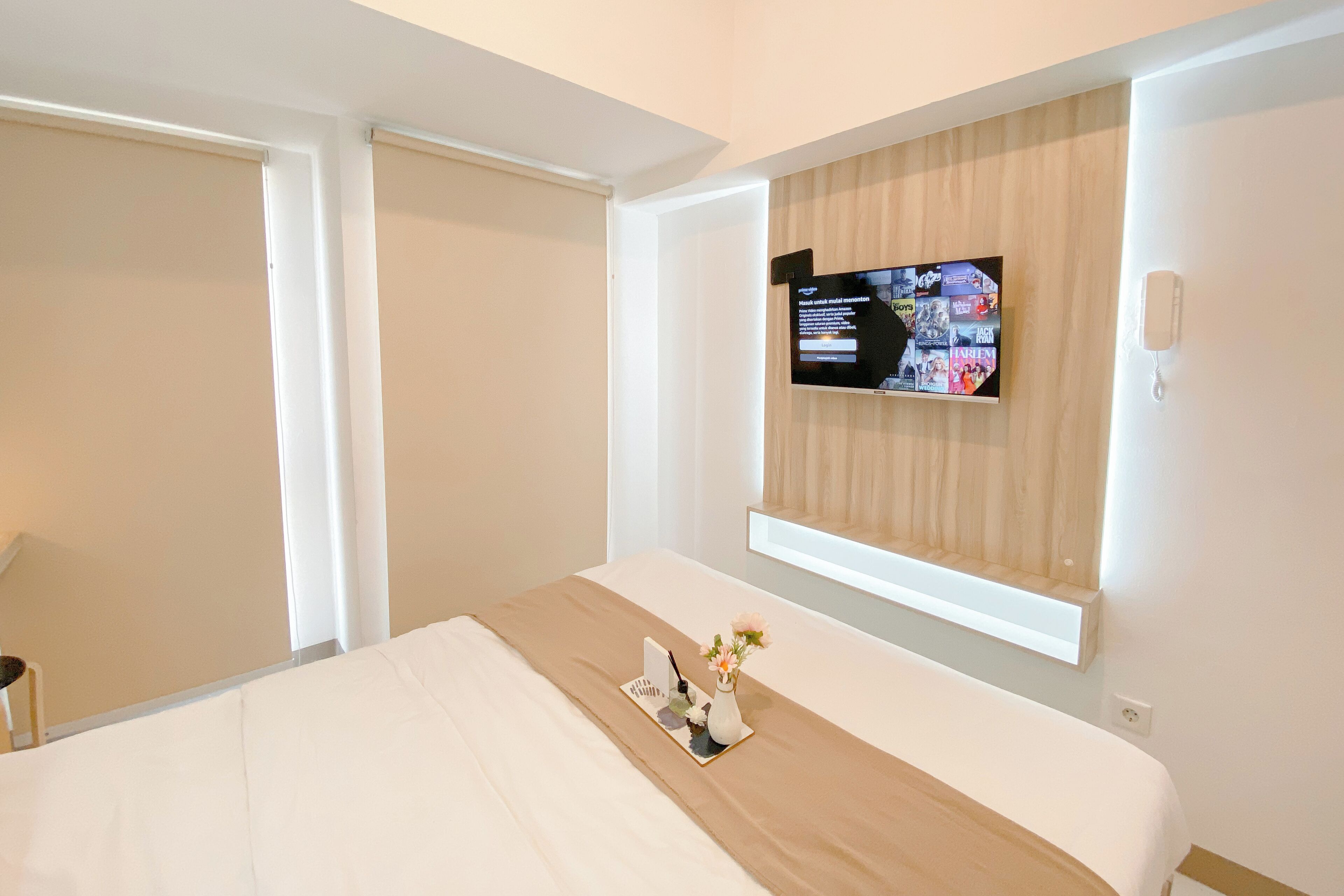 Hotellihuone, jossa on sänky, seinälle asennettu televisio, yöpöytä, jossa on maljakko ja lehti, sekä ikkuna, jossa on sälekaihtimet.
