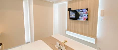 Hotellihuone, jossa on sänky, seinälle asennettu televisio, yöpöytä, jossa on maljakko ja lehti, sekä ikkuna, jossa on sälekaihtimet.