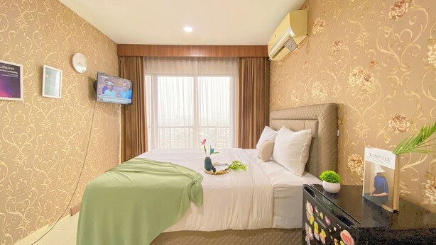 Une chambre d’hôtel comprenant un lit, une télévision, une fenêtre avec des rideaux, une horloge murale et une plante décorative.